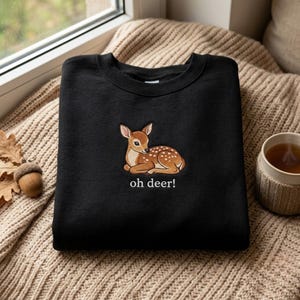 Könnte beinhalten: Ein schwarzes Sweatshirt mit Rundhalsausschnitt und einer Stickerei eines braunen Rehkitzes mit weißen Punkten auf der Brust. Unter dem Rehkitz ist der Text "oh deer!" in weißem Garn gestickt. Das Kleidungsstück hat lange Ärmel und einen Rippkragen.