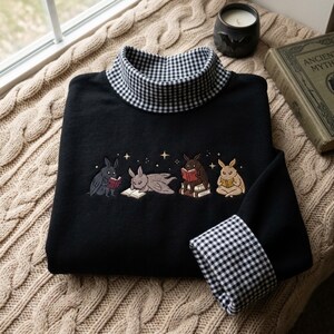 Può includere: Una maglia nera a maniche lunghe con collo alto caratterizzato da un motivo a quadretti vichy bianco e nero. Anche i polsini presentano un motivo a quadretti abbinato. Il petto mostra un ricamo di quattro conigli che leggono libri con stelle.