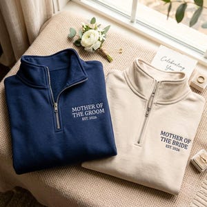 Può includere: Una felpa blu navy con zip a un quarto e testo bianco MOTHER OF THE GROOM EST. 2026 e una felpa beige con testo blu navy MOTHER OF THE BRIDE EST. 2026, entrambe dotate di cerniere in metallo color argento e colletti alti.