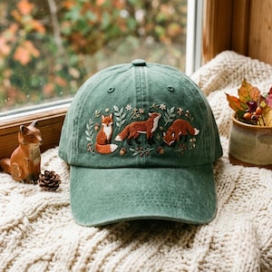 May include: A forest green baseball cap featuring detailed embroidery of three orange foxes surrounded by small white flowers, green leaves, and brown acorns. The cap has a structured crown, a curved brim, and visible stitching across the front panels.