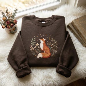 Puede incluir: Una sudadera de cuello redondo color marrón oscuro con un diseño bordado de un zorro sentado dentro de una corona circular de hojas, flores y estrellas. El zorro es de color naranja y blanco, e incluye detalles de una luna creciente y estrellas.