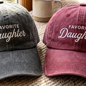 May include: Two baseball caps, one in charcoal gray and one in maroon, both featuring a distressed fabric finish. Each cap has the text "FAVORITE" in a simple sans-serif font and "Daughter" in a white cursive script embroidered on the front panel.