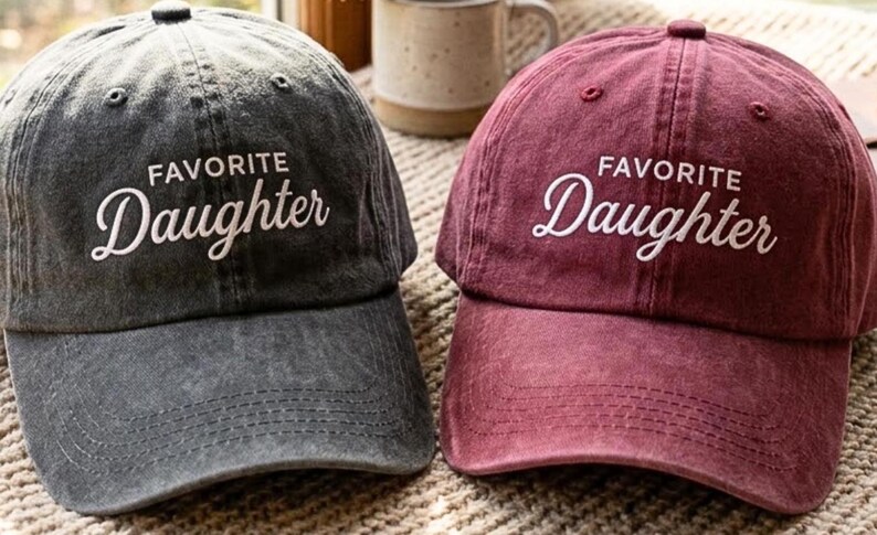 May include: Two baseball caps, one in charcoal gray and one in maroon, both featuring a distressed fabric finish. Each cap has the text "FAVORITE" in a simple sans-serif font and "Daughter" in a white cursive script embroidered on the front panel.