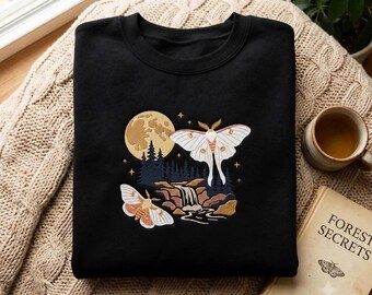 Puede incluir: Una sudadera negra de cuello redondo con un bordado que muestra una polilla blanca grande, una más pequeña, una luna llena, un bosque oscuro y una cascada. El diseño utiliza tonos blancos, tostados y azul oscuro para crear una escena nocturna.
