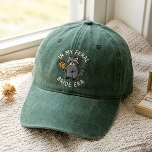 May include: A forest green baseball cap featuring an embroidered graphic of a grey raccoon holding a small bouquet of orange and yellow flowers. The text "IN MY FERAL BRIDE ERA" is stitched in white thread in an arc around the raccoon illustration.