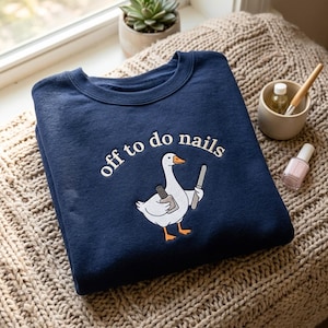 Könnte beinhalten: Ein marineblaues Sweatshirt mit Rundhalsausschnitt und einer Stickerei einer weißen Gans, die eine Nagellackflasche und eine Nagelfeile hält. Der Text "off to do nails" ist in Weiß über der Gans gestickt, die einen orangefarbenen Schnabel hat.