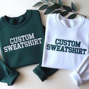 May include: A dark green sweatshirt with white embroidered text reading CUSTOM SWEATSHIRT and a white sweatshirt with dark green embroidered text reading CUSTOM SWEATSHIRT, both featuring a crew neckline and long sleeves with ribbed cuffs.