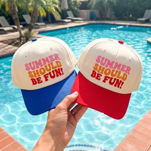 May include: Two baseball caps with off-white crowns and embroidered text that reads SUMMER SHOULD BE FUN! in pink, orange, and red. One cap features a blue brim and blue top button, while the other has a red brim and red top button.