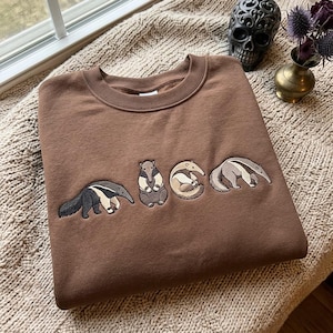 May include: A brown crewneck sweatshirt featuring an embroidered design of four anteaters in various poses across the chest. The embroidery uses shades of grey, cream, and black thread to depict the animals walking, sitting, and curled up.