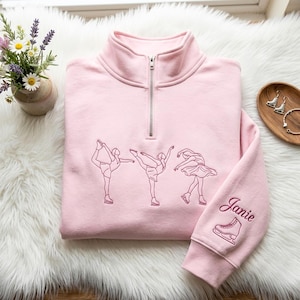 Puede incluir: Una sudadera rosa claro con cremallera de un cuarto y bordados en rosa oscuro de tres patinadores sobre hielo en diferentes poses. La manga izquierda incluye el nombre Janie bordado sobre un pequeño gráfico de un patín.