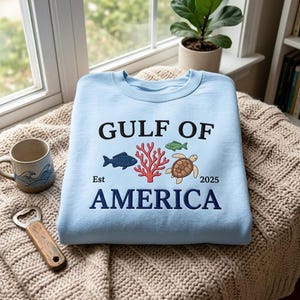 Op de afbeelding: Een lichtblauw sweatshirt met ronde hals en geborduurde tekst GULF OF AMERICA, Est 2025, met een centraal ontwerp van een donkerblauwe vis, een rode koraaltak, een kleine groene vis en een bruine zeeschildpad met een beige schild.