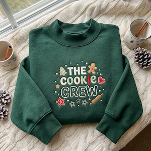 May include: A dark green long-sleeve sweatshirt featuring embroidered text that reads THE COOKIE CREW. The design includes colorful stitched icons of a gingerbread man, a Christmas tree, a heart, stars, and a wooden rolling pin.