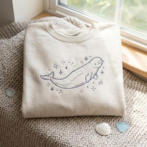 Peut inclure: Un sweat-shirt à col rond de couleur crème avec une broderie bleu foncé représentant un béluga entouré de petites étoiles et de points. La broderie est centrée sur la poitrine du vêtement, qui présente un col et des poignets côtelés.
