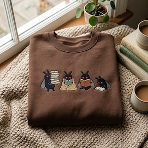Op de afbeelding: Een bruin sweatshirt met ronde hals en een geborduurde afbeelding van vier kleine, zwarte, motachtige wezens met rode ogen. De wezens zijn afgebeeld terwijl ze boeken vasthouden, in een deken gewikkeld lezen, een open boek lezen of op de grond in een boek lezen.