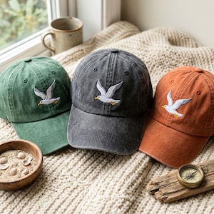 Op de afbeelding: Drie baseballpetten in groen, antracietgrijs en gebrand oranje, elk voorzien van een geborduurde witte meeuw met een gele snavel. De petten hebben een verwassen textuur en zijn gerangschikt op een crèmekleurige, gestructureerde ondergrond.