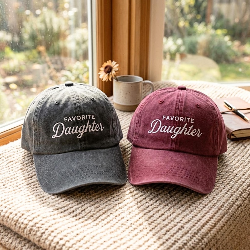 May include: Two baseball caps made of washed cotton fabric, one in charcoal gray and one in maroon. Both caps feature the embroidered white text "FAVORITE Daughter" on the front panel, with a curved brim and a structured crown design.