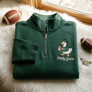 Peut inclure: Un sweat-shirt vert foncé à fermeture éclair quart de tour avec une broderie représentant une oie blanche portant un bonnet et un maillot verts marqués Philly, tenant un ballon de football marron. Le texte Philly Goose est brodé en dessous.