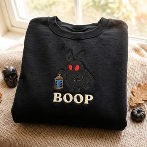 May include: A black crewneck sweatshirt featuring an embroidered design of a fuzzy moth with bright red eyes holding a small lantern. Below the moth, the word BOOP is embroidered in cream-colored capital letters. The fabric has a soft texture.