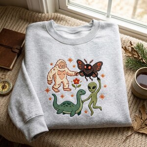May include: A light gray crewneck sweatshirt featuring an embroidered graphic of a beige Bigfoot, a brown moth, a green alien, and a green sea creature holding hands in a circle around a small orange flower, accented by orange stars.