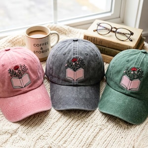Peut inclure: Trois casquettes de baseball en rose, gris anthracite et vert sauge, chacune ornée d'une broderie représentant un livre ouvert avec une fleur rouge et des feuilles vertes sortant des pages. Les casquettes ont une texture de tissu délavé.