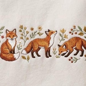 May include: An off-white fabric featuring detailed embroidery of three foxes in various poses, surrounded by small botanical elements including green leaves, yellow flowers, and brown berries, arranged in a horizontal decorative pattern.
