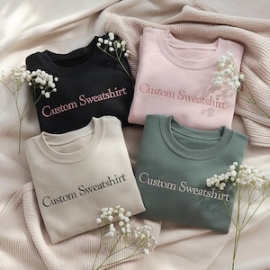Könnte beinhalten: Vier Rundhals-Sweatshirts in Schwarz, Hellrosa, Beige und Salbeigrün. Jedes Kleidungsstück ist auf der Brust mit dem Text "Custom Sweatshirt" in einer kontrastierenden Garnfarbe bestickt. Die Artikel sind auf einer weichen Stoffoberfläche angeordnet.