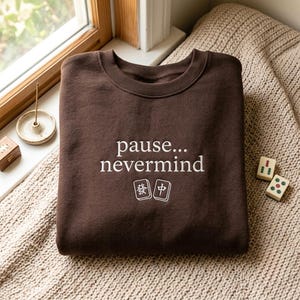 May include: A dark brown crewneck sweatshirt featuring white embroidered text that reads "pause... nevermind" centered on the chest. Below the text are two white embroidered mahjong tiles, one displaying the character for "Fa" and the other for "Zhong".