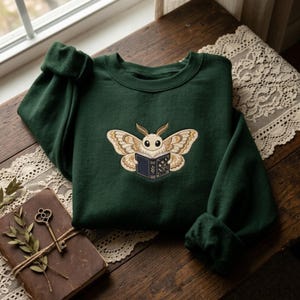 May include: A dark forest green crewneck sweatshirt featuring an embroidered design of a cream-colored moth with large wings holding an open dark blue book with gold floral details and white pages centered on the chest.