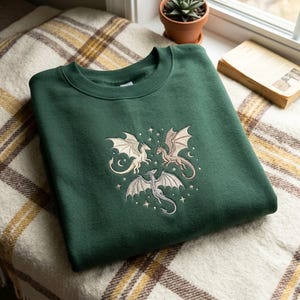 Puede incluir: Una sudadera de cuello redondo en color verde bosque con un diseño bordado de tres dragones de colores claros dispuestos en un patrón circular, rodeados de pequeñas estrellas y lunas crecientes cosidas en el centro de la prenda.