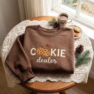 May include: A brown crewneck sweatshirt featuring embroidered text that reads Cookie dealer, with two chocolate chip cookies replacing the letter O and a small red star above the text. The sweatshirt has long sleeves with ribbed cuffs.