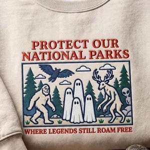 以下が含まれることがあります： 「PROTECT OUR NATIONAL PARKS」および「WHERE LEGENDS STILL ROAM FREE」という文字が刺繍されたベージュのクルーネックトレーナー。森の中にサスクワッチ、幽霊、鹿、鳥、エイリアンが描かれたデザインが施されています。