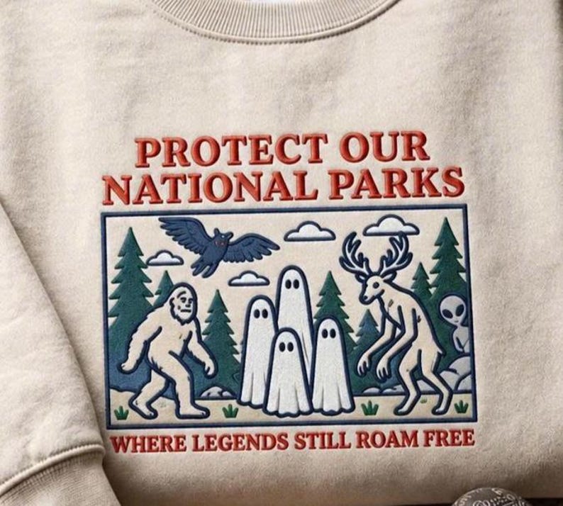 以下が含まれることがあります： 「PROTECT OUR NATIONAL PARKS」および「WHERE LEGENDS STILL ROAM FREE」という文字が刺繍されたベージュのクルーネックトレーナー。森の中にサスクワッチ、幽霊、鹿、鳥、エイリアンが描かれたデザインが施されています。