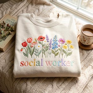 Puede incluir: Una sudadera de cuello redondo en color beige con flores silvestres bordadas en el pecho y el texto "social worker" cosido debajo del diseño floral en una fuente multicolor. La prenda cuenta con mangas largas y un cuello acanalado.