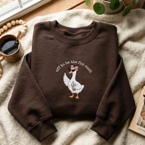 May include: A dark brown crewneck sweatshirt featuring an embroidered graphic of a white goose wearing black sunglasses and a pink bow, holding a coffee cup. The text "off to be the fun aunt" is embroidered in a curved arc above the goose.
