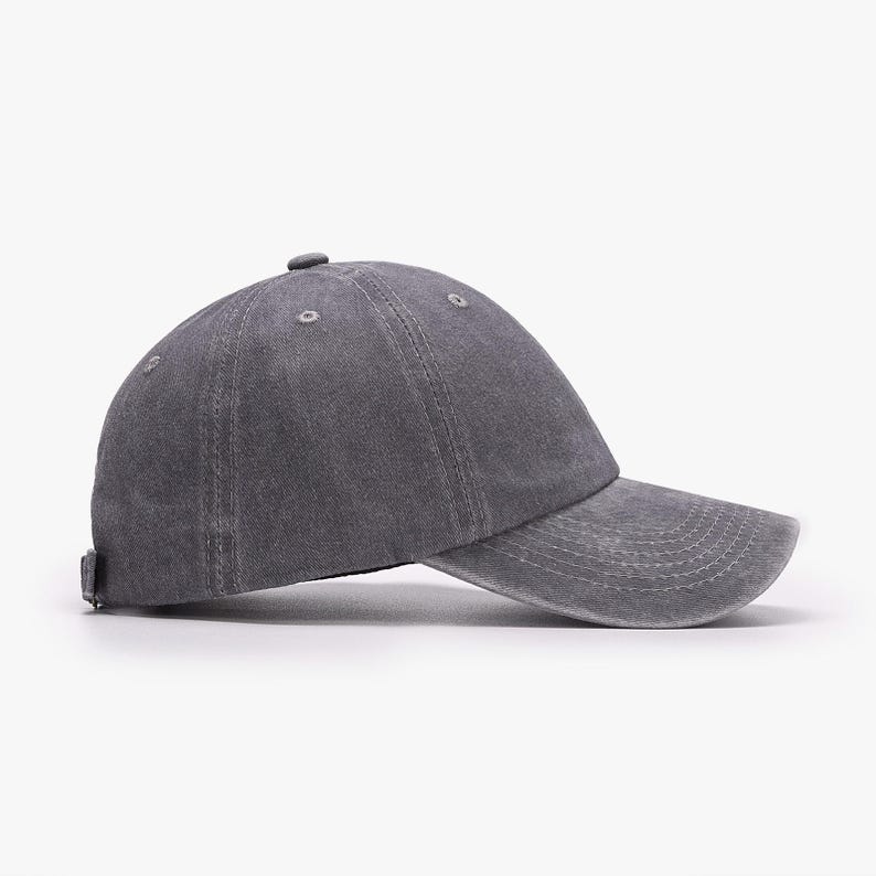 May include: A charcoal gray baseball cap with a washed, distressed fabric finish. The cap features a six-panel construction with stitched eyelets, a curved brim with horizontal stitching, and a small fabric-covered button at the crown center.