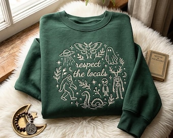 May include: A dark green crewneck sweatshirt featuring a cream-colored graphic print of various cryptids, including a moth, ghost, alien, and Bigfoot, surrounding the text "respect the locals" arranged in a circular pattern with botanical accents.