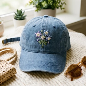 May include: A blue denim baseball cap featuring an embroidered bouquet of purple, white, and light blue columbine flowers with green stems and leaves. The cap has a curved brim, a button on top, and a fabric strap with a metal buckle at the back.
