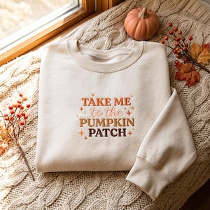 May include: A beige crewneck sweatshirt featuring embroidered text that reads "TAKE ME to the PUMPKIN PATCH" in varying shades of orange and brown, accented with small star and dot symbols centered on the front of the garment.