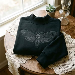 May include: A black long-sleeve sweatshirt featuring a mock neck collar and a detailed embroidered design on the chest. The embroidery depicts a moth with a skull pattern on its thorax, rendered in dark gray thread against the black fabric.