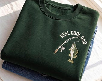 Puede incluir: Una sudadera de cuello redondo en color verde bosque oscuro con un bordado de una caña de pescar y un pez. El texto REEL COOL DAD aparece arqueado sobre la caña en letras blancas. La sudadera está doblada sobre unos vaqueros azules.