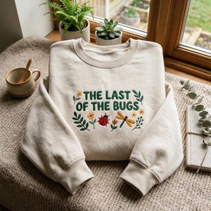 May include: A beige crewneck sweatshirt featuring dark green embroidered text that reads THE LAST OF THE BUGS. The design includes colourful embroidered details of a red ladybug, a yellow dragonfly, small orange flowers, and green leafy stems.