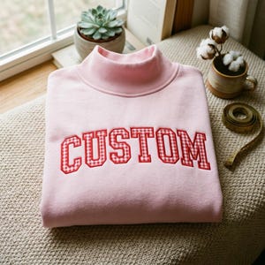Puede incluir: Una sudadera de color rosa claro con cuello alto y la palabra CUSTOM bordada en el pecho con un patrón de cuadros vichy en rojo y blanco. Las letras presentan una tipografía gruesa de estilo bloque con un contorno cosido en rojo.