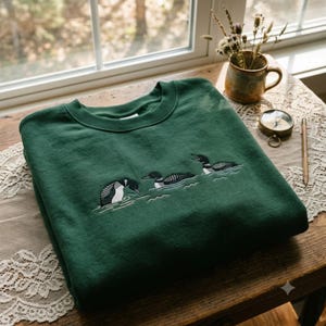 May include: A forest green crewneck sweatshirt featuring an embroidered design of three loons swimming in water. The birds are depicted in black, white, and grey thread, showing detailed plumage and ripples on the water surface below them.