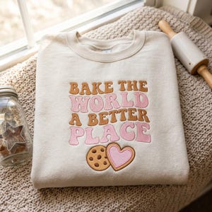 May include: A beige crewneck sweatshirt featuring embroidered text that reads BAKE THE WORLD A BETTER PLACE in alternating brown and pink wavy letters. Below the text is an embroidered brown chocolate chip cookie and a pink heart cookie.