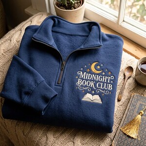 Pode incluir: Uma camisola azul-marinho com fecho de correr curto e um design bordado no peito. O bordado inclui uma lua crescente dourada, pequenas estrelas, o texto "MIDNIGHT BOOK CLUB" em branco e um gráfico de um livro aberto com espirais.