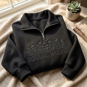 Puede incluir: Una sudadera negra con cremallera de cuarto de largo que presenta un diseño bordado de flores silvestres sobre el texto SOCIAL WORK. La prenda tiene un cuello alto, una cremallera metálica plateada y puños acanalados en las mangas largas.