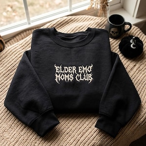 Può includere: Una felpa nera a maniche lunghe con la scritta ELDER EMO MOMS CLUB ricamata in un carattere bianco, stilizzato e frastagliato al centro del petto. Il capo presenta uno scollo rotondo e polsini a coste sulle maniche e sull'orlo inferiore.
