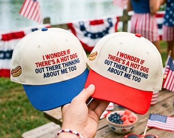 May include: Two baseball caps with off-white crowns and embroidered text that reads I WONDER IF THERE'S A HOT DOG OUT THERE THINKING ABOUT ME TOO. One cap has a blue brim and the other has a red brim, both featuring a small hot dog icon on the side.