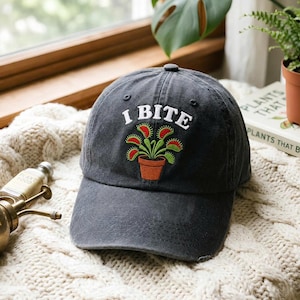 May include: A charcoal grey baseball cap featuring an embroidered graphic of a green Venus flytrap in a terracotta pot. Above the plant, the text I BITE is stitched in white capital letters. The cap has a distressed, washed-out fabric finish.