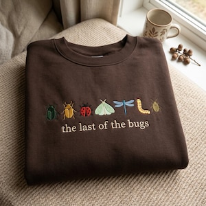 May include: A dark brown crewneck sweatshirt featuring embroidered colorful insects including a green beetle, yellow beetle, red ladybug, light green moth, blue dragonfly, yellow caterpillar, and brown beetle with the text the last of the bugs.
